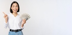 Smiling young modern asian woman, pointing at banner advertisement, holding cash money dollars, standing over white background