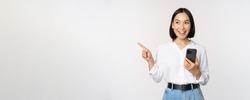 Excited asian girl shopping on mobile app, holding phone and pointing finger left at empty copy space, showing promo text, standing with smartphone against white background