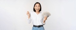 Enthusiastic young asian woman looking excited at camera, holding money dollars in hand, standing over white background