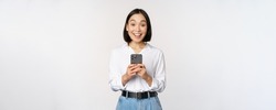 Excited asian woman smiling, reacting to info on mobile phone, holding smartphone and looking happy at camera, standing over white background