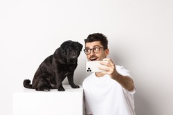 Happy dog owner taking selfie with cute black pug, smiling and looking with love at doggy, holding mobile phone, white background