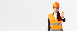 Serious-looking disappointed asian female architect, construction manager at working area wearing safety helmet, showing stop gesture, prohibit action, forbid trespassing, white background