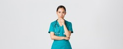 Covid-19, healthcare workers, pandemic concept. Thoughtful female doctor, asian nurse in scrubs looking curious at camera, thinking, searching for solution, standing white background pondering