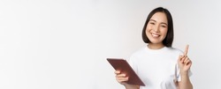 Enthusiastic asian woman with tablet, raising finger and looking amazed, pointing up, standing over white background