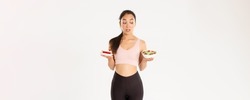Active lifestyle, fitness and wellbeing concept. Portrait of indecisive and tempting cute asian girl trying resist temptation as holding delicious cake, being on diet, looking at healthy salad