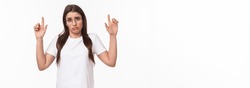 Waist-up portrait of distressed, sad and gloomy indecisive young brunette woman in t-shirt and glasses, shrugging pointing fingers up and making miserable uneasy face, dont know, cant decide.
