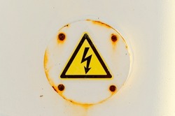 Closeup of electrical risk warning sing with rusty screws on white metallic surface. Triangular shape and black pictogram with yellow background. 