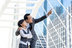 business man wearing a suit embracing his son's shoulder with one arm and pointing a finger for his son to look at a building in the city center with a proud smile because that was his place of work