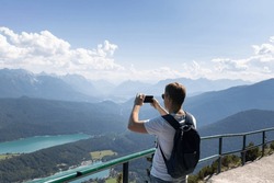 a man with a backpack behind his back photographs a beautiful landscape on his phone. A man standing on an observation deck overlooking mountains and lakes holds a phone in his hands.