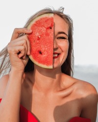 Happy smiling young woman with slice of watermelon half hiding face. Vacation photography.