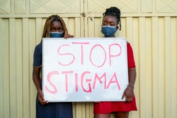 Two beautiful African women wearing locally made mask,looking sad, holding a campaign sign on stop social stigma with covid-19 (corona virus ). Concept on black millennials education on stop stigma