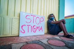 A beautiful young black woman, wearing a locally made mask sitting beside a placard educating on stop stigma on people with corona virus disease - black millennial covid-19 and stigmatisation.