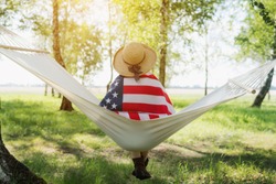 Closeup back view of a Proud woman enjoying summer sunset outdoors and holding american flag. Relax on a hammock in the Park.