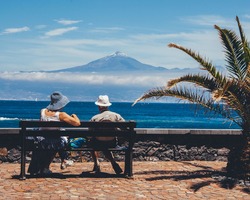 Senior couple enjoying blu skies sea/ocean and mountain view  from island keyside bench in surroundings of a palm. Tourists taking break catching calmness of the sea and restoring the energy.