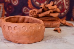 An amateur pinch pot with round circles decorating its rim against a mandala tapestry on parchment paper. Pile of unused terracotta clay in the back, closeup shot in artist workshop.