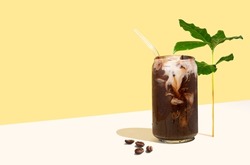 Cold coffee drink, coffee beans and leaves on a yellow isometric diagonal projection background. Can-shaped glass cup of coffee with plant-based milk. Concept of sustainable summer drinks. Creative.