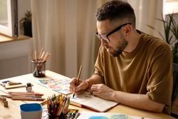 A young artist makes a brush sketch at his desk. Portrait of a creative person in his studio. Creative studio, lifestyle, the process of creating a work of art, the search for inspiration.