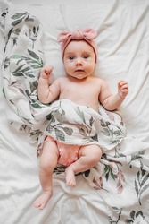 Top vide of cute infant in trendy clothes and bow lying on soft bed under coverlet with floral ornament. Stylish baby with muslin swaddle blanket  on bed