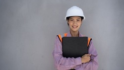 Portrait of a cute looking Asian woman as an engineer, construction inspector, project secretary smiling with a grey wall background.A woman who works smart, smart, confident, hard hat, holding files.