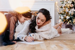 mom and little daughter in the New Year's interior write wishes on a piece of paper. Christmas tree at home. Have a good mood on New Years Eve. Concept preparation to New Year holidays
