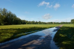 Water floods field path. Puddle on a gray road between green field. Blue sky with clouds.
