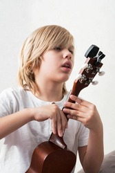 Young boy tuning ukulele at home, close up. Teenage boy sitting on couch playing acoustic guitar.