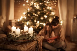 Child sits with mother in front of the Christmas tree and read a book together and look forward to X-Mas, advent wreath