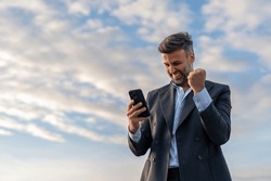 Handsome happy businessman feeling excited raising fist in yes gesture looking at cell phone celebrating work success, getting new approved job opportunity succeed in career and financial goals.