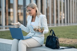 Bothered troubled young blond female business person looking at laptop on break checking emails and eating healthy food. Business woman having discussion talking by conference online virtual chat.