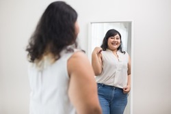 Body positive plus size female clothing. Body positive plus size woman happily and is proud of herself looking at mirror at home. Try wearing clothes that measure the size.