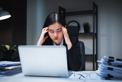 Stressed asian business woman working late at night in the office hands on head feeling headache. Tired woman looking at laptop working hard sitting in the dark room office. Overtime concept