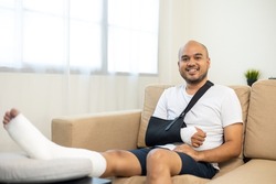 Happy man recovery from accident fracture broken bone injury with leg splints in cast neck splints collar arm splints sling support arm in living room. Social security and health insurance concept.