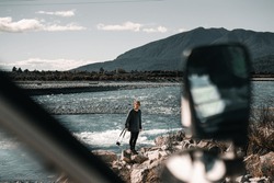 caucasian boy standing on rock with camera strap looking at photographer from shore of shallow river near mountains and forest, taramakau river, new zealand