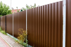 Close-up of a brown metal profile fence. Corrugated surface. Copy space. Security. Private property fencing. Opaque hedge. Outdoor house exterior. Steel material. High. Side view. Urban or indastrial