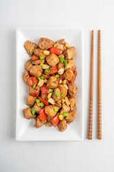 Top view of Kung Pao chicken, also Gong Bao a spicy, stir-fried Chinese dish made with cubes of chicken, peanuts, vegetables served on rectangular plate with chopsticks on white wooden background