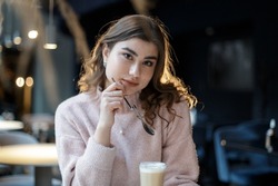 Young beautiful happy woman with long curly hair enjoying latte from a double glass cup, sitting in a cafe.