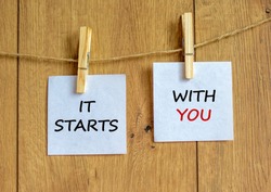 Wooden clothespins with white sheets of paper. Text 'it starts with you'. Beautiful wooden background. Business concept, copy space.