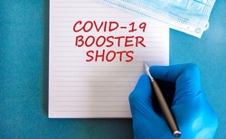 Covid-19 booster shots vaccine symbol. White note with words Covid-19 booster shots, beautiful blue background, doctor hand and metallic pen. Medical mask. Covid-19 booster shots vaccine concept.