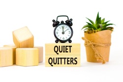 Quiet quitters symbol. Concept words Quiet quitters on wooden blocks. Beautiful white table white background. Black alarm clock. House plant. Business and quiet quitters concept. Copy space.