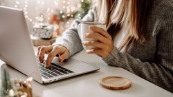 Close up of woman hands with, gifts, coffee cup and laptop. Online shopping at Christmas holidays. Freelance girl woking from home office. Female typing at notebook computer. Christmas moments.