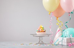 Rainbow Birthday cupcake on a stand with presents, hats and colorful balloons over light grey background. Scene from a birthday party!
