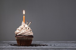 Chocolate Birthday cupcake with  buttercream icing and orange candle over a grey background.