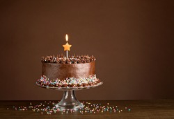 Chocolate birthday cake with sprinkles and candles over a brown background.