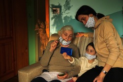 beautiful young little girl granddaughter and her older sister and their old grandmother of Balzac age and each in a handmade medical mask and a beige sweater are sitting watching and reading a mobile