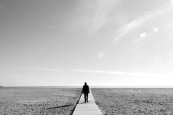 
Imagen en blanco y negro en la que se ve a un hombre caminando por una pasarela de madera sobre la arena de una playa gigantesca
