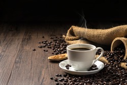 Hot coffee in a white coffee cup and many coffee beans placed around and sugar on a wooden table in a warm, light atmosphere, on dark background, with copy space.