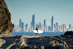Surfer walking along a rocky coastline with the Gold Coast city skyline in the background.