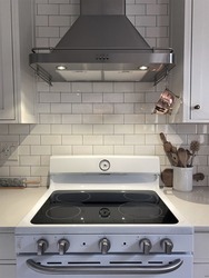 White Kitchen cupboard with white subway tile. Kitchen utensils are in a bowl ready for cooking with a small vintage copper colander hanging above the stove with a large kitchen hood exhaust.