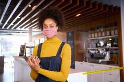 Female African coffee shop owner wearing face mask standing in front of counter with digital tablet.
