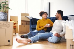 Young mixed ethnicity couple taking a break on moving day into new home sitting on floor in lounge drinking coffee surrounded by removal boxes
